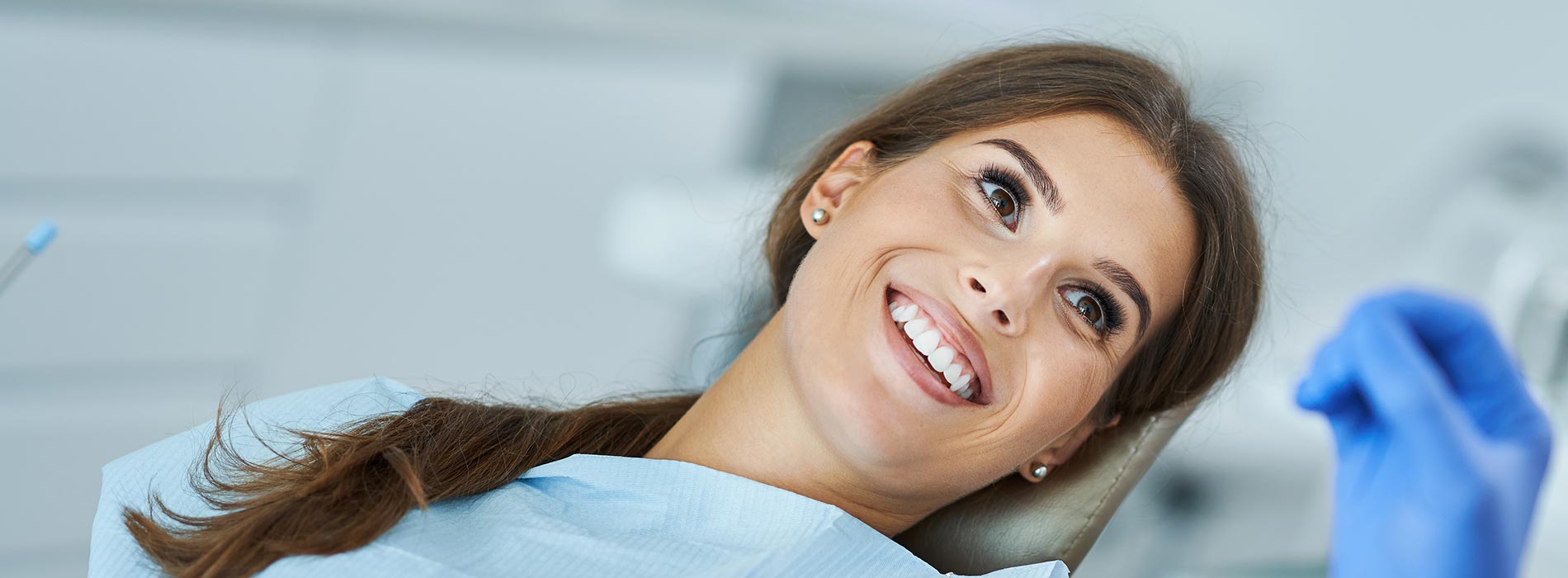A woman with blue eyes and a smile, wearing a white lab coat and a stethoscope around her neck, gazes directly at the camera while seated in a dental chair.
