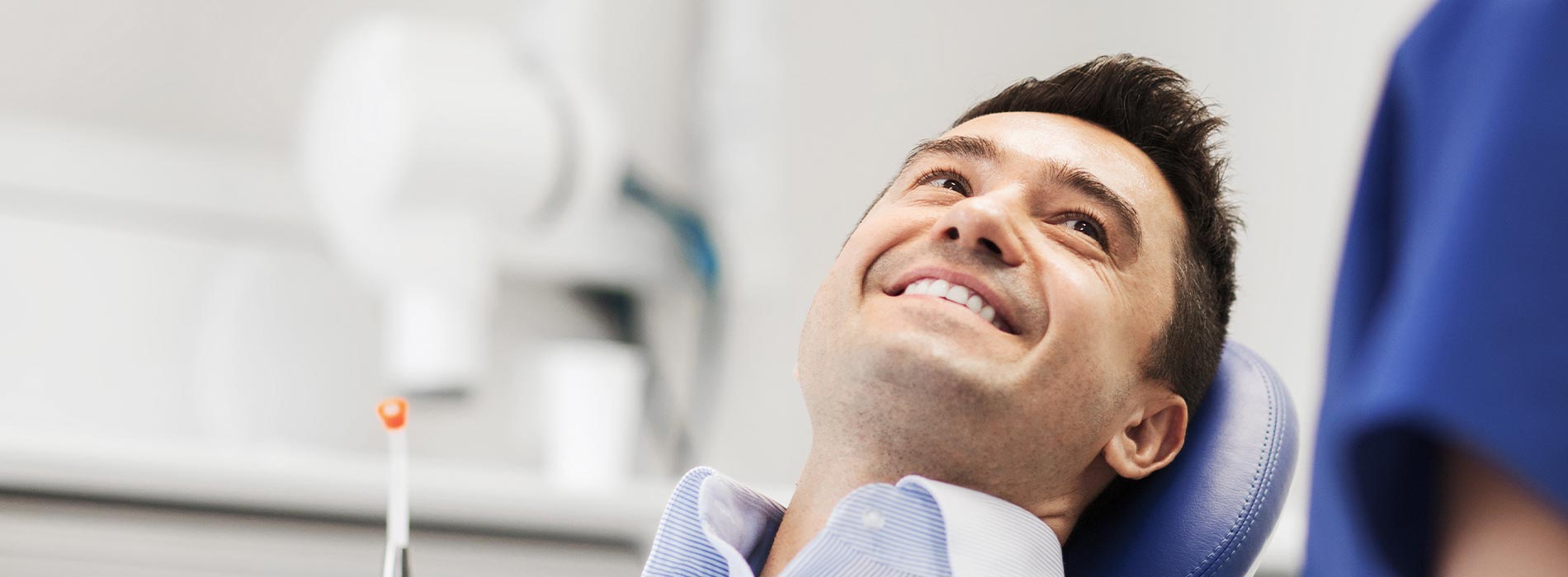 The image shows a man sitting in a dental chair with a smile on his face, being attended to by a dental professional who appears to be smiling as well.